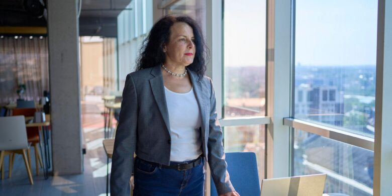 Woman in her 60s in business attire working at a laptop by an office window, considering Medicare options before turning 65